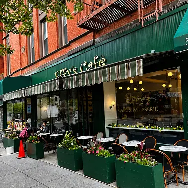 A cozy café with green awnings, outdoor seating, and potted plants, located on a street corner.