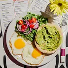A plate with avocado toast, fried eggs, and a side salad on a table with menus and a flower.
