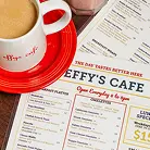 A red saucer holds a white coffee cup with a menu for Effy's Cafe in the background.